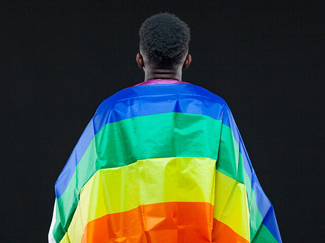 gettyimages1285870288-640x480-1 Back view of young african american man wrapped in a rainbow flag isolated on black backgr